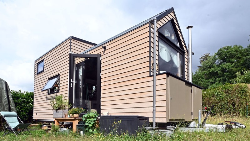 Ein Tiny House aus Holz steht auf der Wiese eines Campingplatzes. Im Hintergrund ist eine Hecke zu sehen.