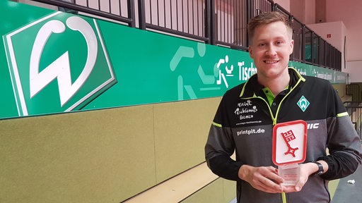Werders Tischtennis-Spieler Mattias Falck steht lächelnd vor einem Werder-Logo mit seiner Auszeichnung zu Bremens Sportler des Jahres in der Hand.