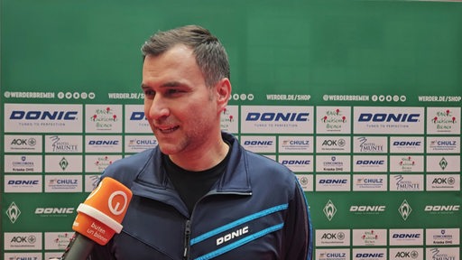 Werders Tischtennis-Trainer Cristian Tamas steht lächelnd vor einer Werbewand beim Interview.