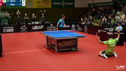 Werders Tischtennis-Profi Marcelo Aguirre sinkt im Siegerjubel auf die Knie und reißt die Arme hoch, die Fans in der Halle feiern ihn.