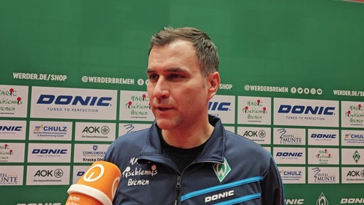 Werders Tischtennis-Trainer Cristian Tamas steht nach dem Spiel vor einer Werbewand beim Interview.