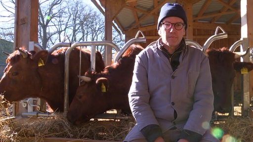 Der Landwirt Tobias Bertzbach sitzt auf Heu, im Hintergurnd seine Kühe. 