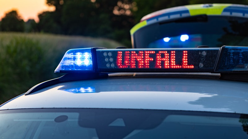 Ein Streifenwagen der Polizei steht mit Blaulicht und dem Schriftzug Unfall im Display an einem Unfallort.