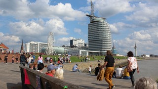 Personen laufen am Wasser entlang. Im Hintergrund ist das Sail City Hotel in Bremerhaven zu sehen.