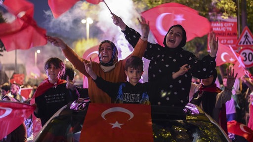 Menschen, darunter Frauen mit Kopftuch, halten Flaggen der Türkei und heben die Arme voller Freude.