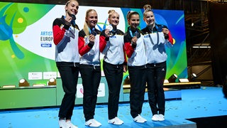 Die Turnerin Karina Schönmaier steht strahlend auf dem Siegerpodest bei der EM neben ihren Teamkolleginnen Helen Kevric, Lea Marie Quaas, Janoah Mueller und Silja Stoehr, die alle ihre Silbermedaillen hochhalten.