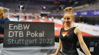 Turnerin Karina Schönmaier steht lächelnd in der Stuttgarter Sporthalle vor dem Hinweisschild zum DTB-Pokal.