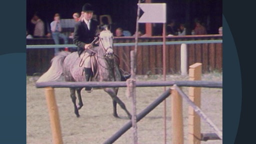 Eine Reiterin auf ihrem Pony bei einem Turnier. Sie reiten auf eine Hürde zu.