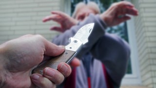 Symbolbild für einen Überfall: Jemand zückt ein Messer, ein Mann kreuzt zum Schutz seine Arme.