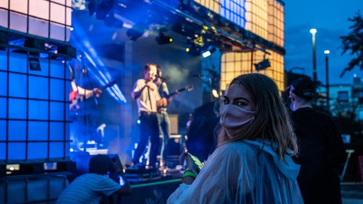 Junge Frau mit Mund-Nasen-Schutz blickt vor Open-Air-Konzert in Kamera