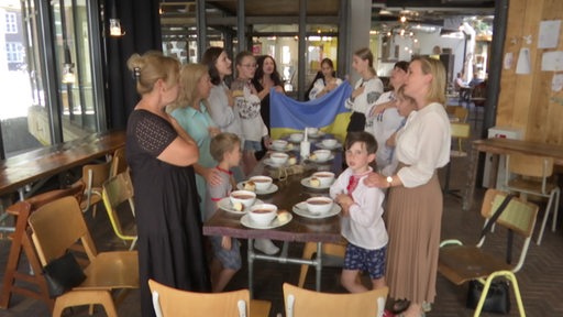 Mehrere Personen stehen mit einer Ukraine-Flagge um einen Tisch mit Essen und singen.