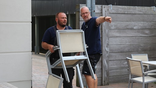 Ruslan Moroz trägt zwei Gartenstühle, sein Kollege Lars Fröhlich zeigt ihm den Weg
