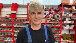 Ein Mann in blauer Latzhose steht in einem Werkzeuglager und schaut in die Kamera