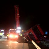 Ein Lkw liegt umgekippt auf der Mittelleitplanke einer Autobahn, im Hintergrund sind ein Kran und Rettungsfahrzeuge zu sehen.