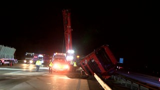 Ein Lkw liegt umgekippt auf der Mittelleitplanke einer Autobahn, im Hintergrund sind ein Kran und Rettungsfahrzeuge zu sehen.