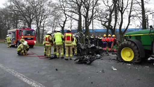 Ein völlig zerstörter VW Touran nach einem Unfall mit einem Traktor, daneben Feuerwehrleute