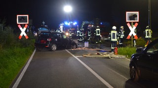 Ein dunkles vorne zusammengestauchtes Auto steht auf einer Landstraße an einem Bahnübergang. Darum stehen zahlreiche Feuerwehrleute, Lichter blinken.