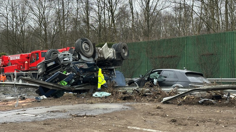 Ein zerstörter Lkw liegt auf dem Dach. Ein kaputtes Auto steht davor. Die Leitplanken sind durchbrochen.