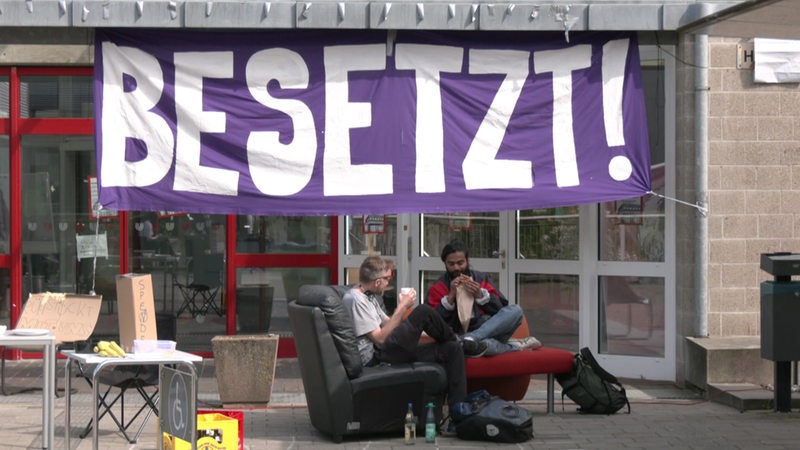Ein großes Banner vor dem Eingang des hörsaals der Uni Bremen auf dem "Besetzt!" steht. Davor sitzen Studierende auf Sesseln.