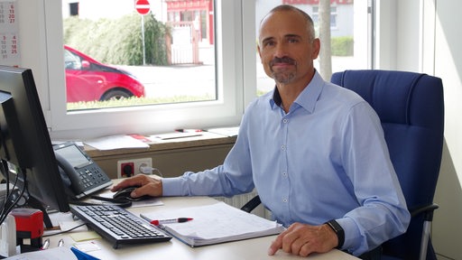 Ein mittelalter Mann mit hellblauem Hemd sitzt im Büro vor einem Computer.