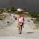 Ein Tourist läuft mit seiner Tochter auf dem Arm aus einem Feriengebiet auf Rhodos. Im Hintergrund rauchschwarzer Himmel