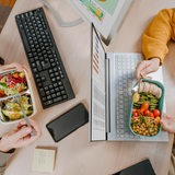 2 Leute sitzen sich an Laptops gegenüber und essen veganes Frühstück aus ihren Boxen.
