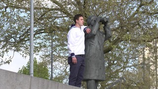 Ein junger Mann steht als Matrose verkleidet neben einer Statue in Vegesack.