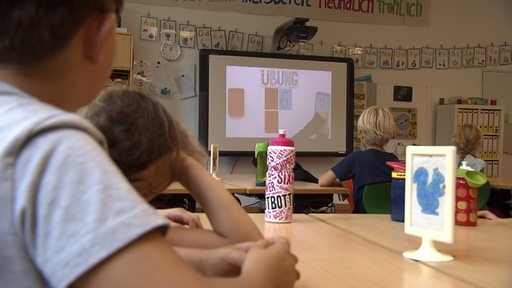 Schülerinnen und Schüler schauen in einem Klassenzimmer auf einen Bildschirm.