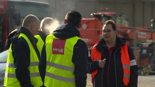 Personen in verdi-Warnweste unterhalten sich und rauchen dabei.