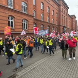 Menschen laufen, teils mit Warnwesten gekleidet, mit Fahnen und Transparenten eine Straße entlang.