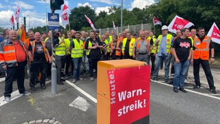 Zahlreiche Menschen mit Westen und Plakaten stehen auf einem Hafengelände.