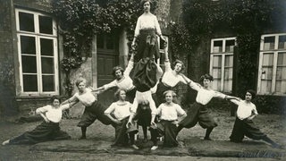 Frauen des Turnvereins Verein Vorwärts in der zweiten Hälfte des 19. Jahrhunderts in weißen Blusen und langen Röcken bilden vor einem Haus eine Pyramide.