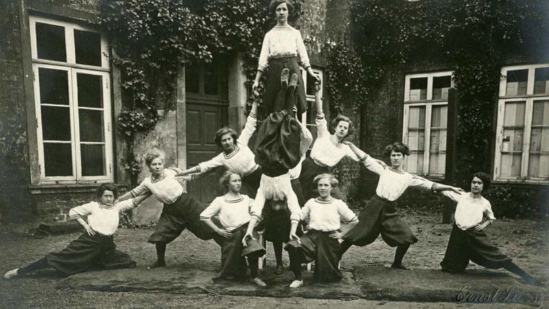 Frauen des Turnvereins Verein Vorwärts in der zweiten Hälfte des 19. Jahrhunderts in weißen Blusen und langen Röcken bilden vor einem Haus eine Pyramide.