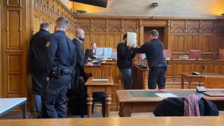 Mehrere Menschen stehen in einem Gerichtsaal, ein Mann hält sich einen Ordner vor das Gesicht.