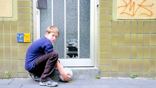Ein Junge mit Ball sitzt an der eingeschossenen Glasscheibe einer Haustür.