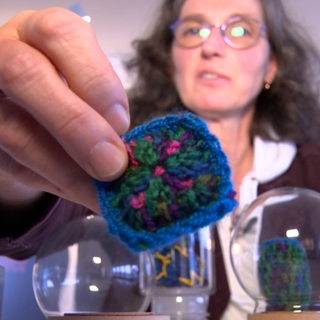 Eine Frau mit Brille hält einen gehäkelten Virus in die Kamera