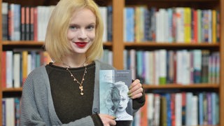 Blonde, stark geschminkte Frau, Mitte 20, vor Bücherregal hält Buch in die Kamera