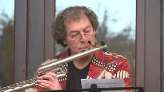 Der Komponist Volker Klein spielt ein Blasinstrument und guckt auf ein Notenblatt. Er steht vor einem Fenster.
