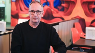 Ein Mann mit Brille in schwarzem Pullover, der Schriftsteller Volker Kutscher, sitzt an einem Tisch.