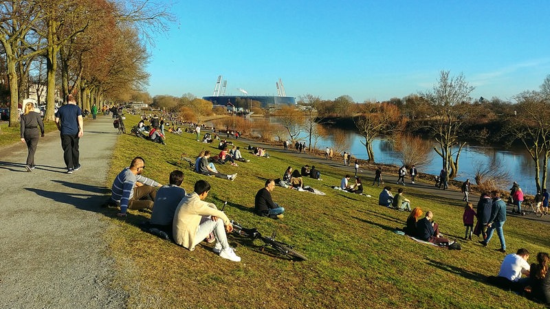 Menschen sitzen an einem Hang am Fluss, im Hintergrund ein Stadion.