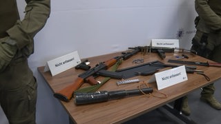 Ein Tisch auf dem sich mehrere Waffen befinden, neben den Waffen stehen Schilder mit der Aufschrift "Nicht anfassen".