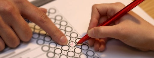 Ein Finger zeigt auf einem Stimmzettel einer anderen Hand, wo ein Kreuz gemacht werden soll. (Symbolbild)