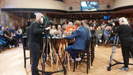Mehrere Menschen an einem Tisch, darum Fernsehkameras