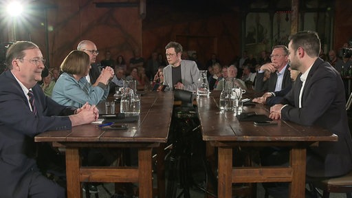 Moderator Felix Krömer und 6 Bremische Spitzenkandidaten sitzen an einem Tisch beim Wahlmobil in Hemelingen.