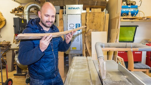 Ein Mann in einer Werkstatt hält ein Stück Holz in der Hand.