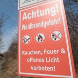 Auf einem Schild steht: "Achtung! Waldbrandgefahr!" Im Hintergrund ist ein Waldstück zu sehen.