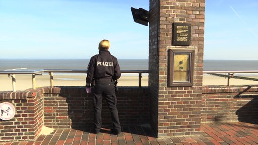 Eine Polizistin steht an einer Mauer. Hinter der Mauer ist der Meeresstrand auf den sie schaut.