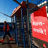 Ein Schild mit der Aufschrift "Heute Warnstreik!" hängt am Zaun neben einem Tor-Eingang, Mann läuft dadurch