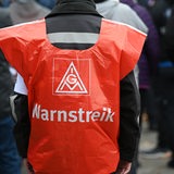 Ein Mensch trägt ein rotes Cape mit dem Logo der Gewerkschaft IG Metall, auf dem "Warnstreik" steht