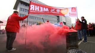 Mehrere Menschen mit Schildern in der Hand stehen während eines Warnstreiks vor einer Tonne mit rotem Rauch.